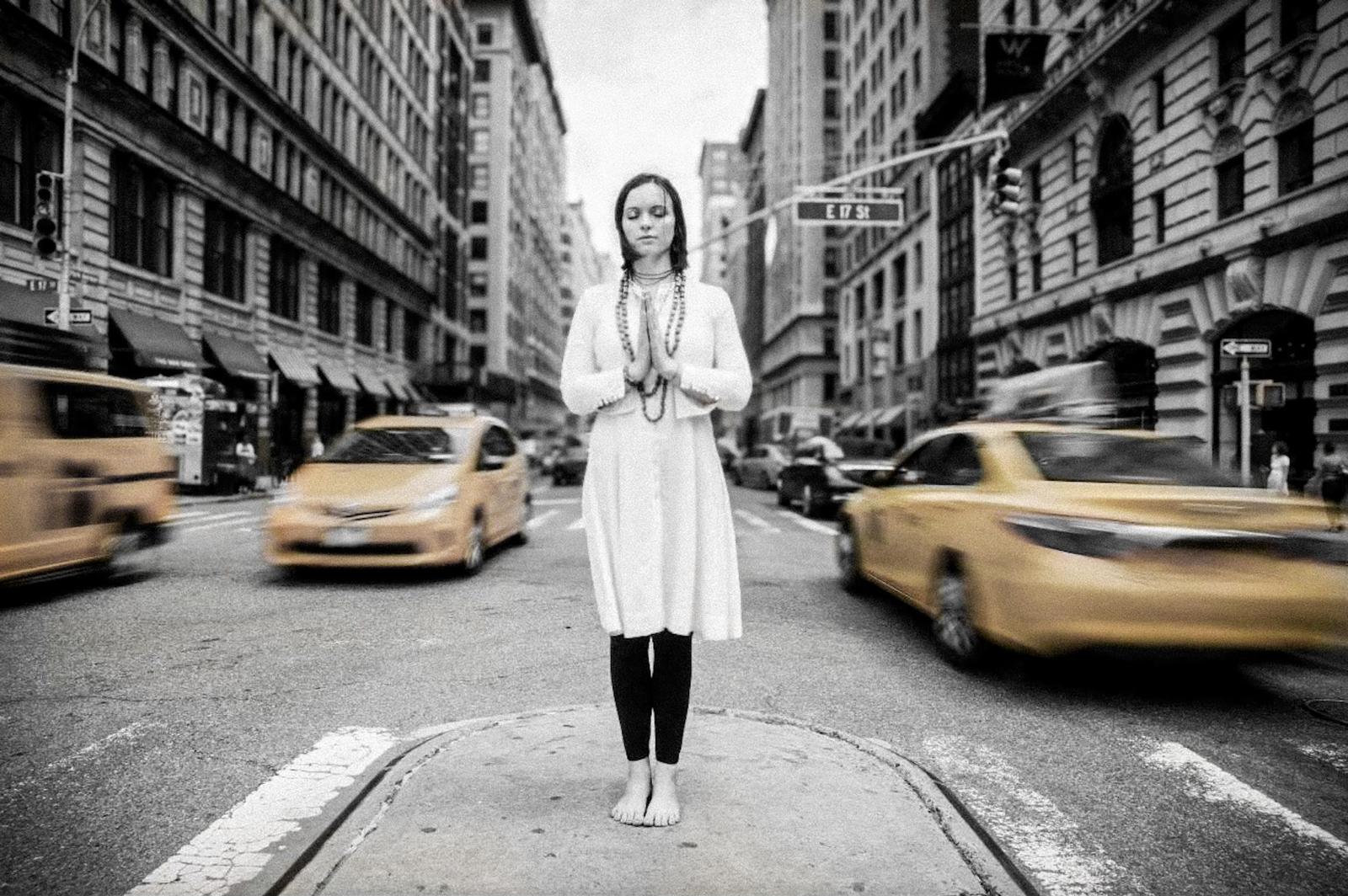 A woman meditating, eyes closed and palms together, standing still among the taxis of a busy New York street