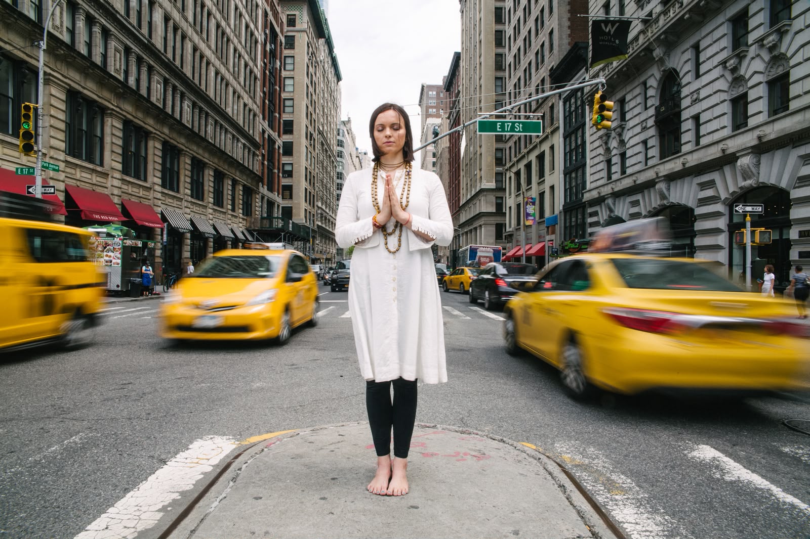 A woman meditating, eyes closed and palms together, standing still in the middle of a busy New York street
